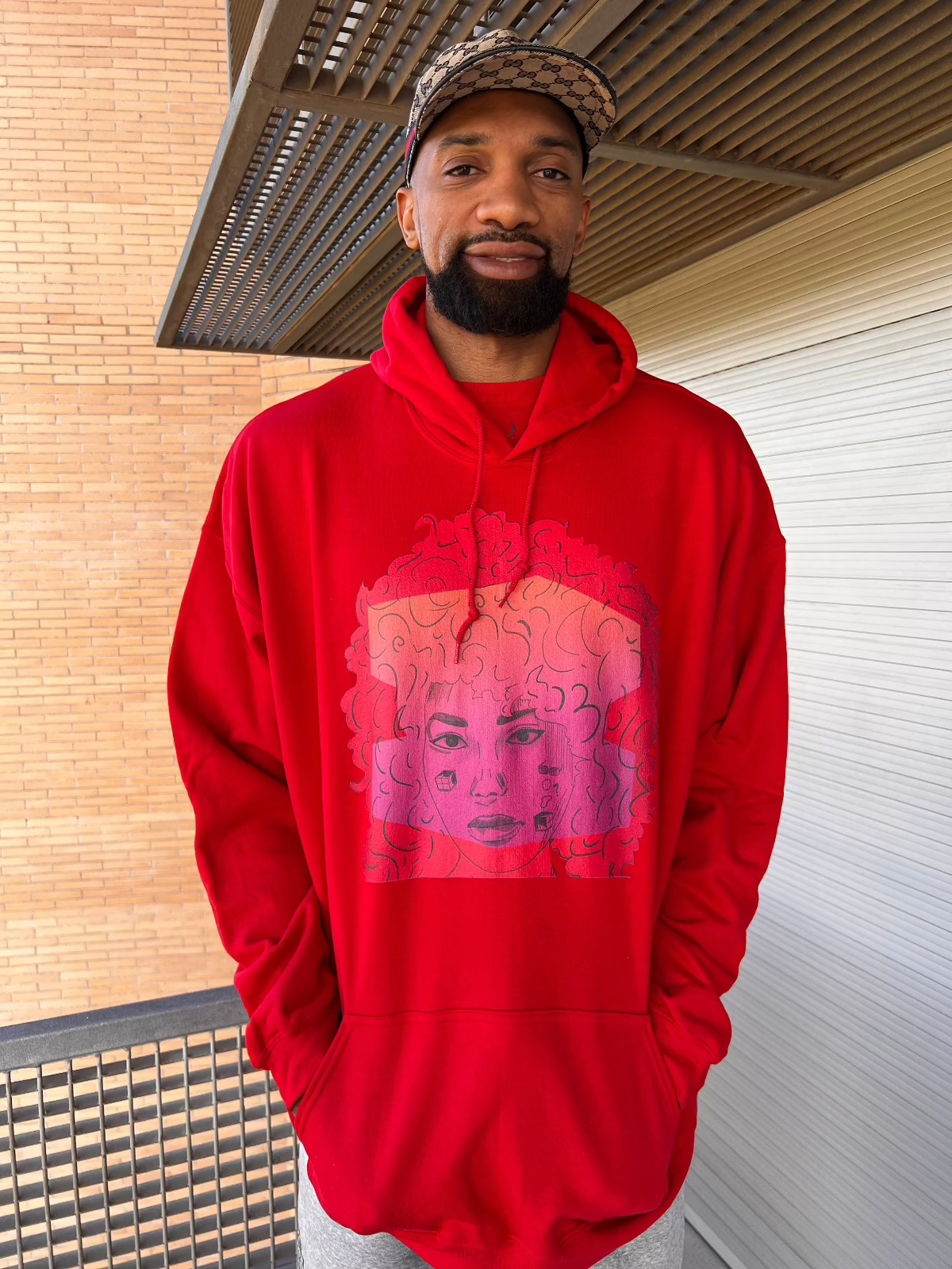 Khem Birch models a unisex red zip-up hoodie featuring Salome Saliya’s face with curly hair on the front, artistic graphic design, geometric accents, and deep red background.