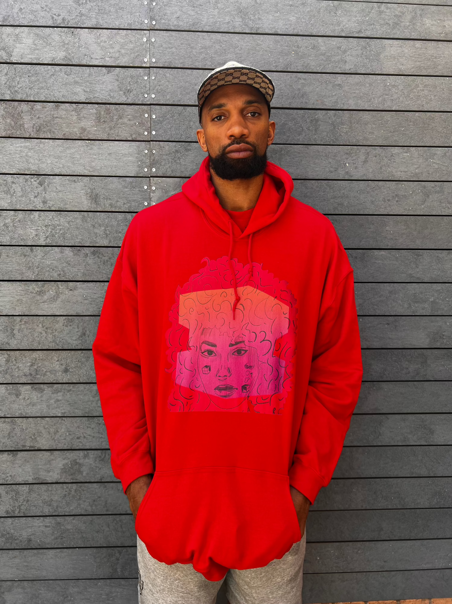 Khem Birch wearing a unisex red zip-up hoodie featuring a bold graphic of Salome Saliya’s face with curly hair on the front, set against a deep red background with geometric line accents.