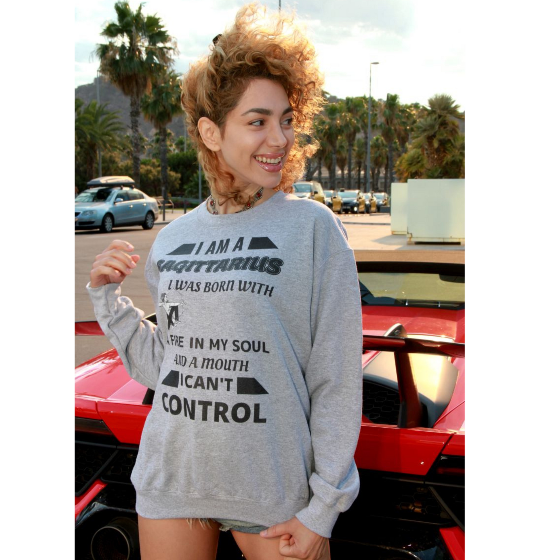 Woman wearing grey Sagittarius zodiac sweatshirt with bold quote – I was born with fire in my soul and a mouth I can’t control – standing in front of red sports car, casual astrology-themed fashion.