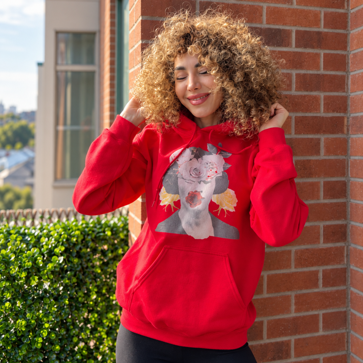 Model with hands in her hair wearing a red graphic hoodie featuring a woman’s portrait decorated with roses.